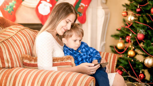 年轻的母亲与小男孩坐在客厅扶手椅上的肖像和在智能手机上观看视频