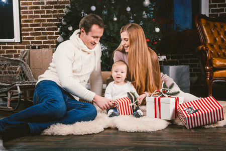 家庭庆祝圣诞晚会。家庭三白种人坐在针叶树圣诞树下的地毯羊毛地板上。孩子拿着礼物盒子, 父母年轻时髦穿戴