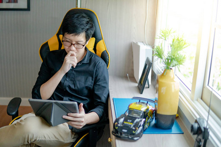 年轻的亚洲商人, 戴着眼镜, 坐在卧室的工作椅上使用数码平板电脑。现代电子小工具理念下的家居生活 lifestlyle