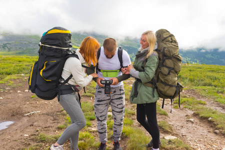 三名游客朋友 一个男人和两个女孩, 金发碧眼, 背上背着背包的红头发, 站在山和云的背景上, 微笑着看着相机上的照片。