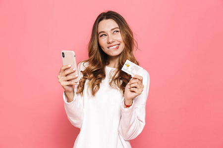 一个欢快的年轻女子的肖像使用手机, 持有塑料信用卡隔离在粉红色背景