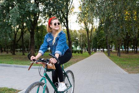 时尚, 快乐的女孩在时尚的衣服和太阳镜骑自行车在公园里, 看着侧身和微笑。在公园里走自行车。活跃在其余的概念