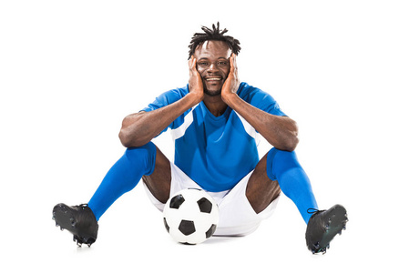 快乐的年轻非洲裔美国足球运动员坐在与球和微笑在相机隔绝白色