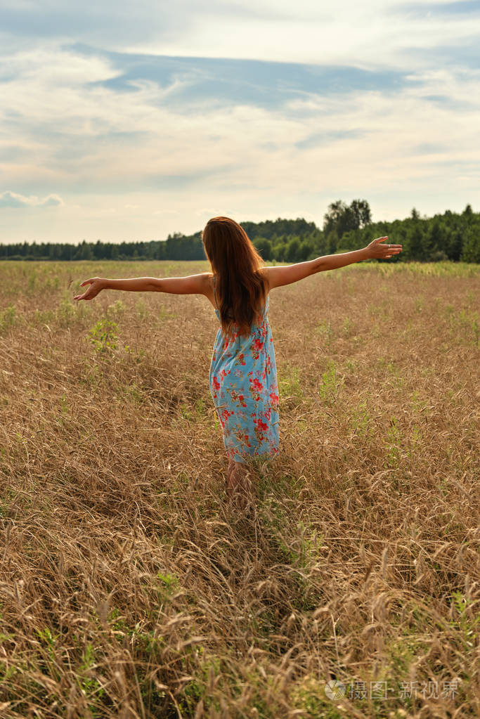 年轻女子女孩站在她的背部张开双臂到日落的麦田在农村
