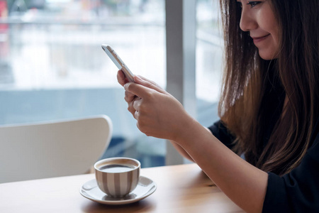 一个美丽的亚洲女人的特写图像手持和使用智能手机与咖啡杯在咖啡馆的木桌上
