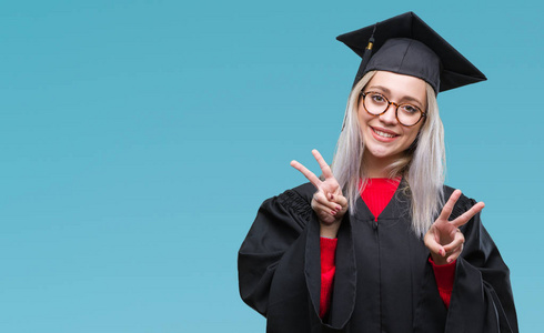 年轻的金发女子穿着毕业制服在孤立的背景下, 微笑着看着相机, 显示手指做胜利标志。第二个
