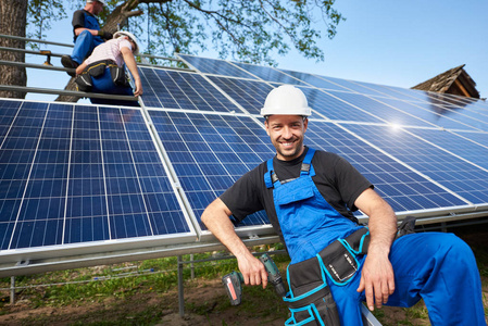 在未完成的高外部太阳能电池板光电系统前, 与高平台上的工人团队一起使用电动螺丝刀的快乐技术员肖像