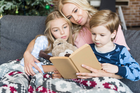 快乐的年轻母亲在圣诞节时在睡衣上看书与可爱的孩子