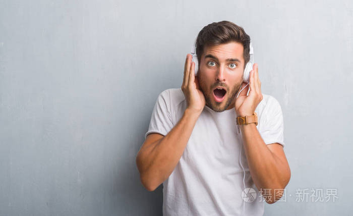 英俊的年轻人在灰色垃圾墙上戴着耳机听音乐吓坏了惊讶的脸, 害怕和