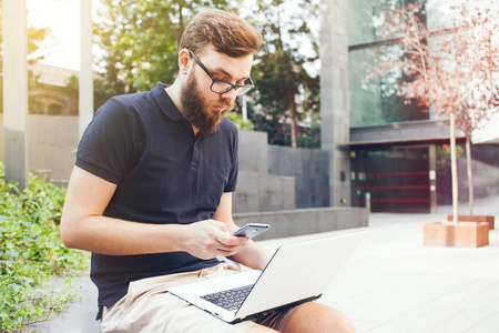 时髦的胡子的年轻人正在户外便携式电脑坐在城市广场