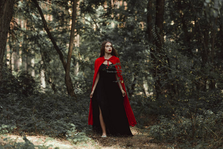 神秘女孩在黑色礼服和红色斗篷漫步在森林里