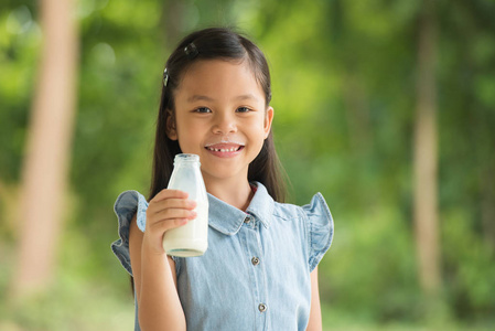 快乐的小孩子, 亚洲女孩黑色的头发在蓝色西装微笑, 享受健康早餐和喝牛奶从瓶在绿色自然背景