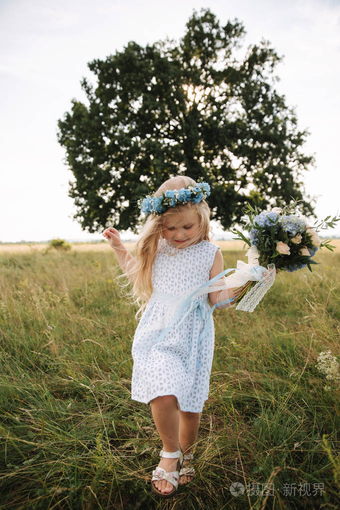 小女孩在天空蓝色礼服与花圈和花束立场在领域在大树前面