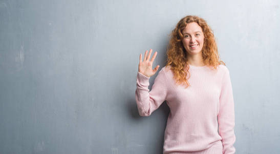 灰色垃圾墙上的年轻红头发的女人穿着粉红色的毛衣显示和指向五的手指, 同时微笑着自信和快乐