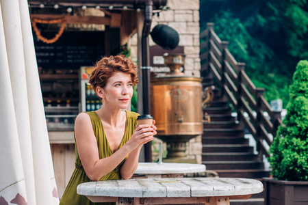 一个穿着绿色连衣裙的漂亮红头发的女人站着, 手里拿着咖啡, 微笑着看着她。夏天, 放松, 假期概念