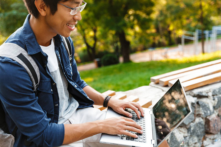 在公园长椅上坐着膝上型电脑的亚洲男性学生微笑的侧面视图