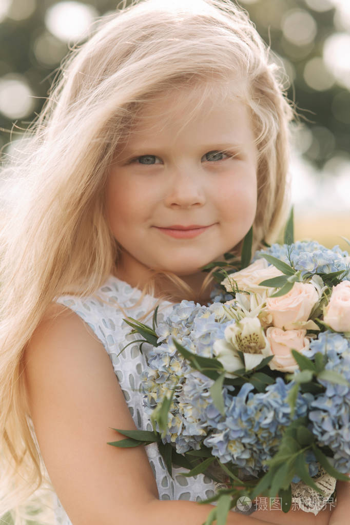 小女孩在天空蓝色礼服与花束站立在领域在大树前面.孩子的微笑和乐趣