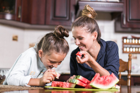 可爱的小女孩和她美丽的妈妈正在切水果, 红色西瓜和微笑, 而在厨房做饭在家里。幸福的家庭