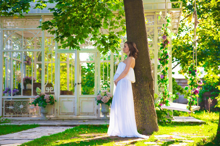 孕妇在自然草坪上与凉亭与花白色礼服