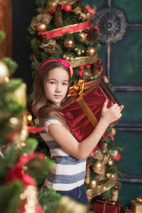 可爱的小女孩，穿着红色头带持有附近圣诞树礼物