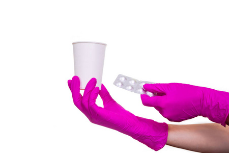 女性手在紫色的医疗手套拿着一个白纸杯和药丸。手臂与药物剂量和水隔离在白色背景