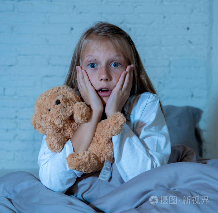 害怕的小女孩坐在床上, 用双手抱着她的泰迪熊捂着她的脸, 害怕在黑暗