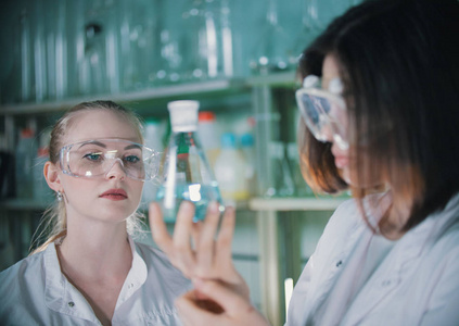 化学实验室里的两名年轻女子拿着一个装有透明液体的烧瓶。金发碧眼的女人在焦点