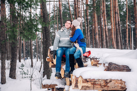 年轻的情侣穿着蓝色毛衣坐在柴火上, 拥抱在冬天的森林里