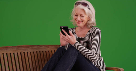 微笑的老白人女性使用移动设备在公园板凳上绿色屏幕