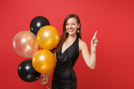 微笑的女孩穿着黑色小礼服庆祝, 指着食指一边, 拿着气球孤立在红色的背景。情人节, 新年快乐生日模拟节日派对概念