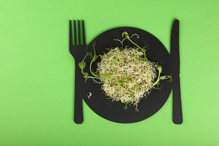 关闭新鲜绿豆和青豆微绿色沙拉芽在黑色的盘子, 叉子和刀形板在绿色背景与复制空间, 高顶视图, 直接上面