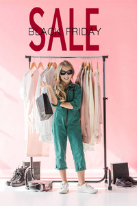 时尚的孩子在工作服和太阳镜站立与购物袋附近衣服和鞋子在精品, 黑色星期五销售横幅概念