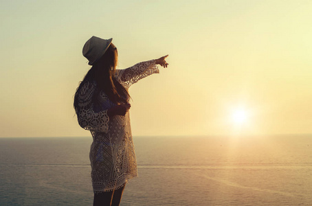 年轻女子在帽子和可爱的夏天衣服站在石头上，越过大海看着日落