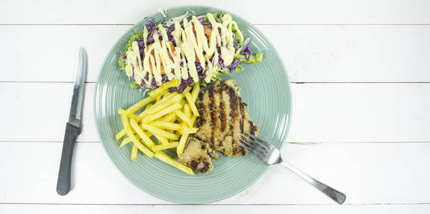 猪肉牛排在一盘白色木地板桌道具装饰叉子和勺子, 薯条, 绿色沙拉, 紫色花椰菜, 顶部视图