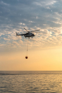 一架消防直升机将水收集到海里, 飞向群山, 在美丽的黎明天空下熄灭森林大火。