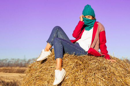 户外大气生活方式照片年轻美丽的女士棕色头发和眼睛在粉红色的外套, 针织帽子和牛仔裤摆姿势和坐在蓝天周围的干草。温暖的秋天
