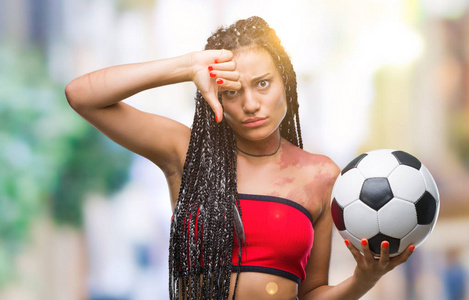 年轻的辫子头发非洲裔美国人与出生标记举行足球在孤立的背景与愤怒的脸, 消极的迹象显示不喜欢大拇指下来, 拒绝的概念