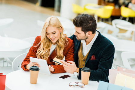 微笑的夫妇在网上购物与信用卡和数字平板电脑, 而坐在咖啡馆在商场