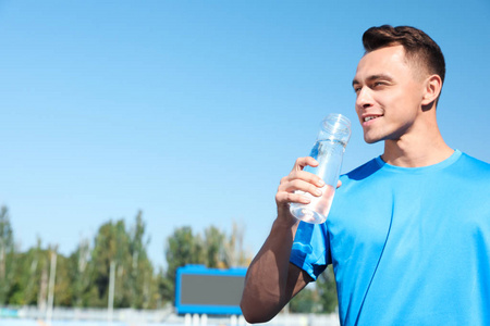 阳光明媚的日子里, 年轻的运动男子在体育场用瓶子喝水。文本的空间