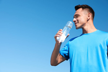 在阳光明媚的日子里, 年轻的运动男子从瓶子里喝水, 对着蓝天