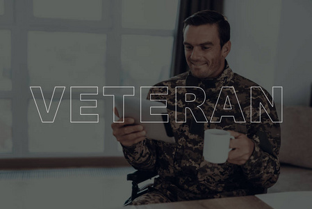 退伍军人。坐在轮椅上的残疾人。在平板电脑上观看视频的人是士兵。身穿军服的士兵在杯中喝咖啡的人。男人的微笑。位于客厅里的男人