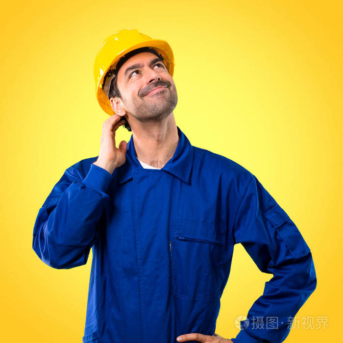 年轻的工人, 带着头盔站着思考一个想法, 同时在黄色背景上抓头