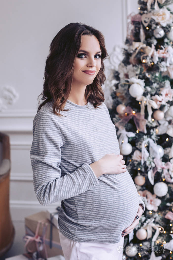 怀孕的美丽女孩手牵着肚子美丽的圣诞树与灯光