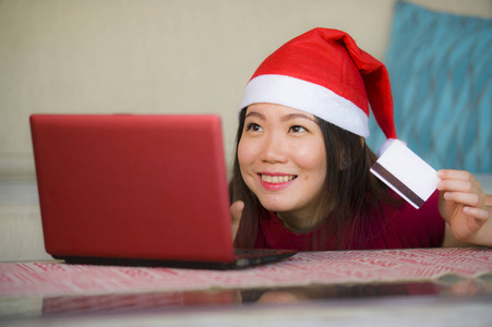 年轻美丽和快乐的亚洲中国妇女在圣诞节圣诞老人帽子拿着信用卡使用笔记本电脑上网购物微笑着兴奋在家里的沙发客厅