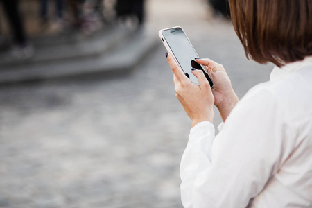 一个美丽的女人站在模糊的街道背景时使用手机的肖像。女孩使用应用程序在她的智能手机
