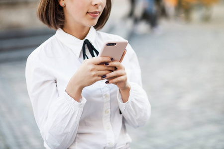 一个美丽的女人站在模糊的街道背景时使用手机的肖像。女孩使用应用程序在她的智能手机