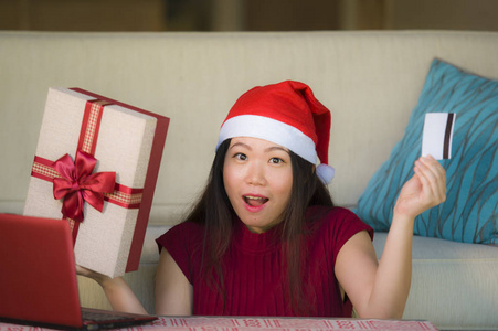 年轻美丽快乐的亚洲中国妇女拿着圣诞老人的帽子和圣诞礼物盒使用笔记本电脑上网购物微笑愉快在家里