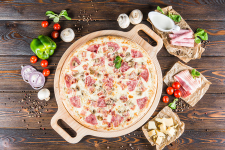 大比萨饼与火腿和蘑菇在一个圆形的切菜板上的一个黑暗的木制背景。披萨食材。顶视图