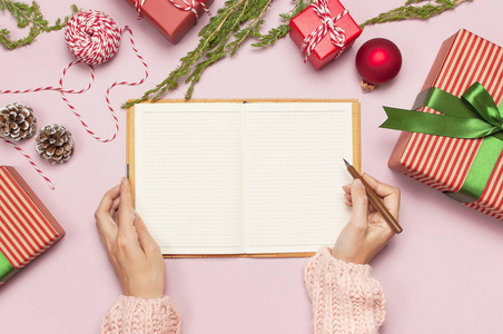 女性手在粉红色的针织毛衣是写在干净的笔记本计划为新的一年, 礼品盒冷杉树枝上粉红色的背景平躺。圣诞规划概念假日装饰