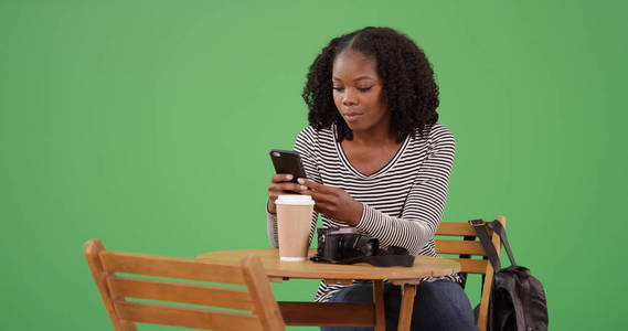 年轻的现代黑人妇女用手机坐在绿色屏幕上的咖啡馆桌旁
